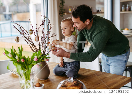 Father and little son decorating pussy willows branches, putting easter eggs on them. 124295302