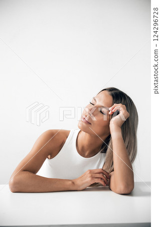 Woman in white tank top rests her head on hand while smiling softly in minimalistic interior Woman in white tank top rests her head on hand while smiling softly in minimalistic interior 124296728