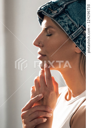 Woman in stylish denim hat gently touching her face while basking in natural light indoors 124296738
