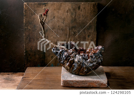 Handmade ceramic bowl with natural branch sprouting from center displayed on stone base against rustic wooden background. Artistic pottery showcasing organic textures and earthy colors. Handmade ceramic bowl with natural branch sprouting from center displayed on stone base against rustic wooden background. Artistic pottery showcasing organic textures and earthy colors. 124298105