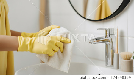 Side-angle close-up of a housewife's hands in yellow gloves cleaning a bathroom sink with a white cloth 124298399