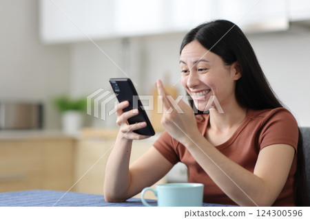 Happy asian woman crossing fingers checking smart phone Happy asian woman crossing fingers checking smart phone 124300596