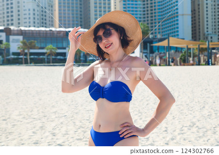 A woman in a blue swimsuit and straw hat stands on the stunning beach of Dubai, gazing at the impressive skyline filled with modern skyscrapers, while the golden sand and gentle waves create a perfect A woman in a blue swimsuit and straw hat stands on the stunning beach of Dubai, gazing at the impressive skyline filled with modern skyscrapers, while the golden sand and gentle waves create a perfect 124302766
