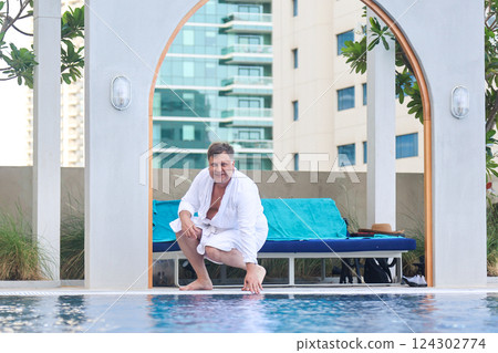 A Man in a White Robe Stands by the Hotel Pool, Preparing to Dive into the Refreshing Water, Embracing a Day of Leisure and Comfort Under the Bright Sun, Ready to Unwind and Enjoy His Vacation 124302774