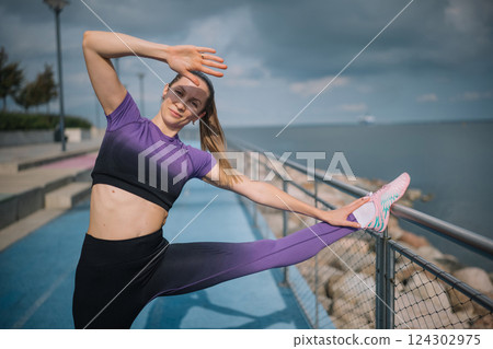 A fitness enthusiast stretching by the waterfront on a cloudy day in a vibrant athletic outfit 124302975