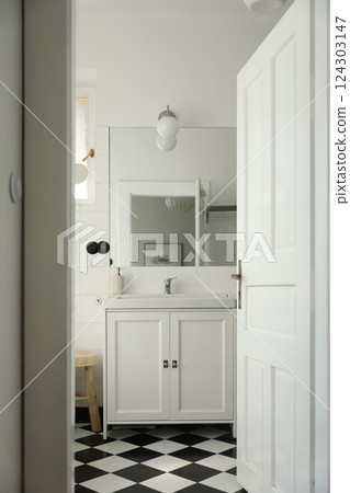 Vintage elegant bathroom interior through open door. Wash basin, cabinet, mirror, glass pendant. Black white checkered floor, white tile wall background. Elegant minimal Scandinavian style design 124303147