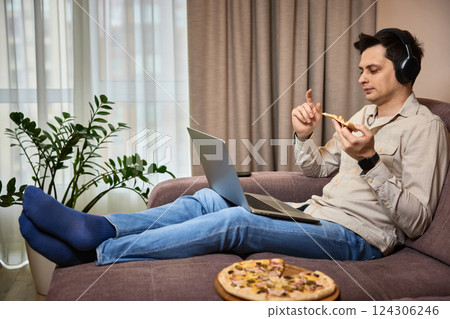 man sitting on sofa and eating pizza while working on laptop 124306246