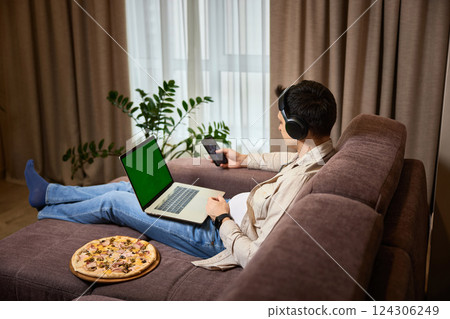 Young man sitting on sofa and eating pizza while working on laptop 124306249