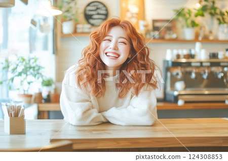 Cheerful woman with curly red hair smiles warmly while leaning on a wooden counter in a bright cafe during daytime 124308853