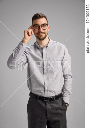 Portrait of young man adjusting glasses, wearing shirt and black pants, looking with confidence and little smile against grey studio background Portrait of young man adjusting glasses, wearing shirt and black pants, looking with confidence and little smile against grey studio background 124309151