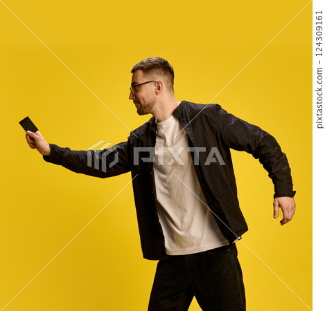 Online payment. Smiling young man in black and white outfit using credit card for convenient payment against yellow studio background 124309161