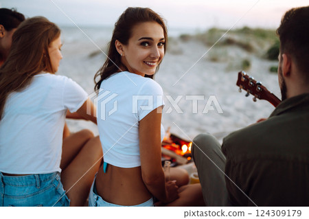 Group of young friends sitting on beach and fry sausages. One man is playing guitar. Camping time. 124309179