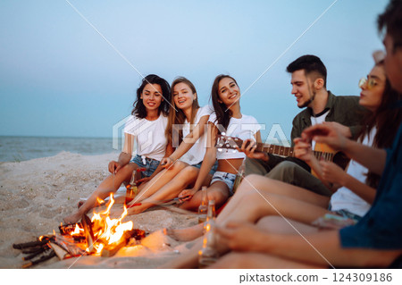 Group of young friends sitting on beach and fry sausages. One man is playing guitar. Camping time. 124309186