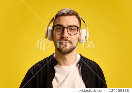 Portrait of young handsome man in black and white outfit, in glasses, listening to music in headphones against yellow background 124309188