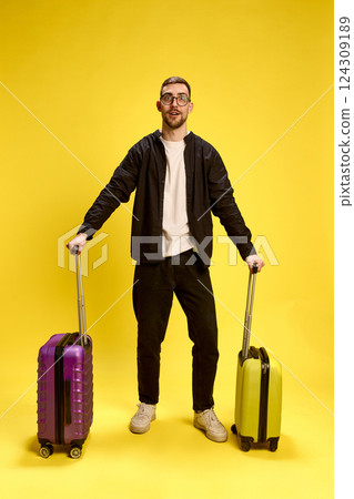 Excited young man dressed in comfortable black outfit, standing with two suitcases in hands and expressing excitement 124309189