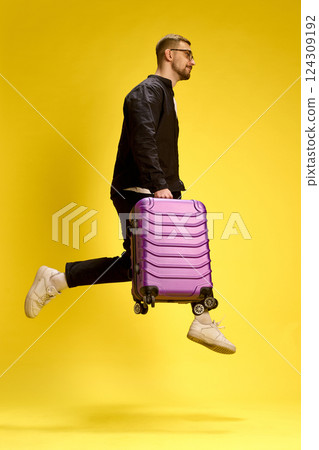 Dynamic man jumping mid-air with suitcase, dressed in black, playful motion on yellow studio background. Dynamic man jumping mid-air with suitcase, dressed in black, playful motion on yellow studio background. 124309192