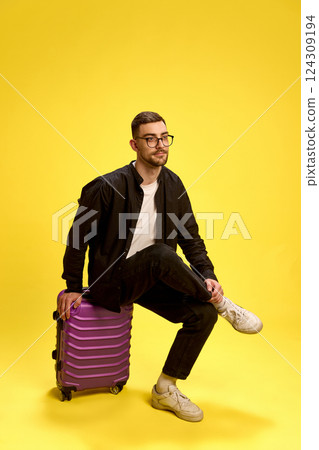 Trendy young man sitting on purple suitcase with confident look, wearing casual black outfit against yellow background. 124309194