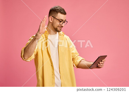 Man in yellow shirt smiling and waving hand while looking at tablet against pink background. Man in yellow shirt smiling and waving hand while looking at tablet against pink background. 124309201