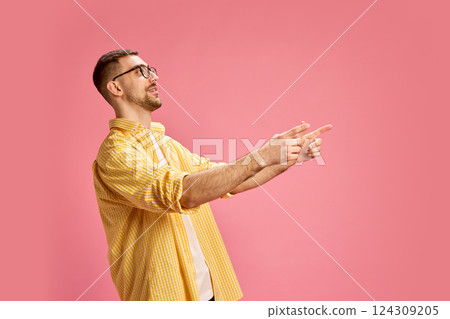 Young man in yellow checkered shirt smiling and pointing fingers forward against pink background. Young man in yellow checkered shirt smiling and pointing fingers forward against pink background. 124309205