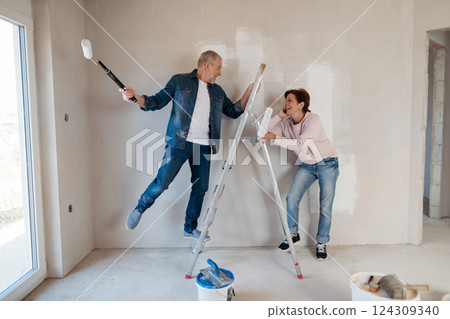 Senior husband and wife standing on a ladder repainting the walls. 124309340