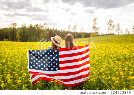 Two Young woman with american flag on blooming meadow. 4th of July. USA flag fluttering in the wind. 124313579