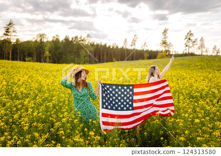 Two Young woman with american flag on blooming meadow. 4th of July. USA flag fluttering in the wind. Two Young woman with american flag on blooming meadow. 4th of July. USA flag fluttering in the wind. 124313580