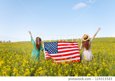 Two Young woman with american flag on blooming meadow. 4th of July. USA flag fluttering in the wind. Two Young woman with american flag on blooming meadow. 4th of July. USA flag fluttering in the wind. 124313583