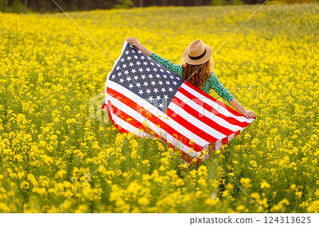 Young woman with american flag on blooming meadow. Independence Day. USA flag fluttering in the wind 124313625