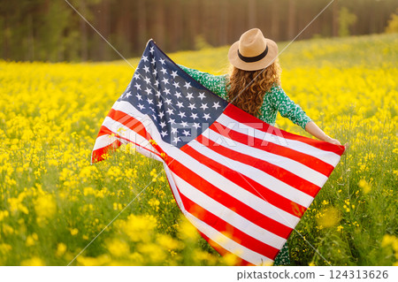 Young woman with american flag on blooming meadow. Independence Day. USA flag fluttering in the wind 124313626