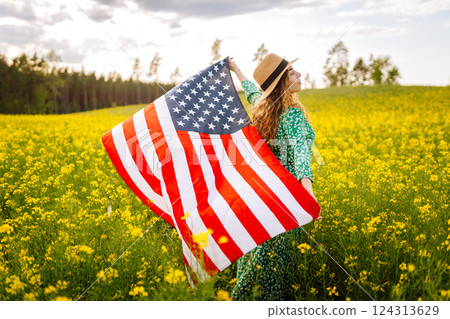 Young woman with american flag on blooming meadow. Independence Day. USA flag fluttering in the wind Young woman with american flag on blooming meadow. Independence Day. USA flag fluttering in the wind 124313629
