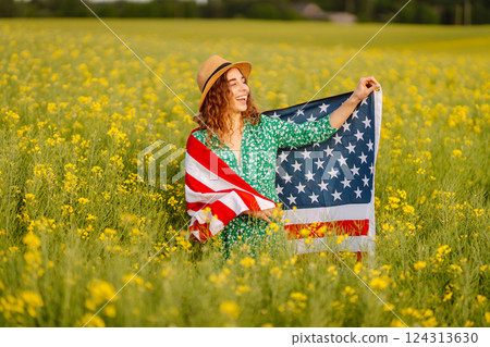 Young woman with american flag on blooming meadow. Independence Day. USA flag fluttering in the wind Young woman with american flag on blooming meadow. Independence Day. USA flag fluttering in the wind 124313630