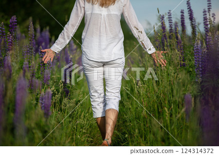 A woman walks with her back to the camera through a field of purple lupine flowers. She is dressed in white and enjoys the beauty of the flowers. 124314372