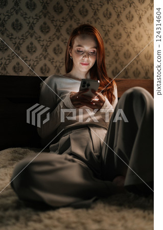 Vertical portrait of focused tired woman using smartphone sitting on bed in dark bedroom at night, light from screen illuminating face. Concept of social media addiction, phone overuse, insomnia. Vertical portrait of focused tired woman using smartphone sitting on bed in dark bedroom at night, light from screen illuminating face. Concept of social media addiction, phone overuse, insomnia. 124314604