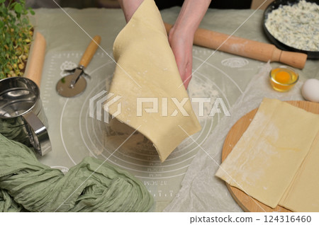 Woman's hands holding a sheet of dough. Rolling pin, cutter, eggs, green towel, microgreens sprouts on kitchen table. Home cooking baking puffs Woman's hands holding a sheet of dough. Rolling pin, cutter, eggs, green towel, microgreens sprouts on kitchen table. Home cooking baking puffs 124316460