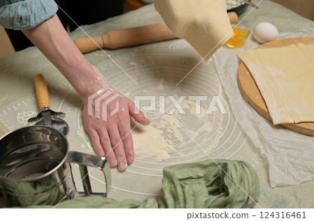 Woman's hand holding a sheet of dough and sprinkles flour worktop. Rolling pin, cutter, eggs, green towel on kitchen table. Home cooking baking puffs Woman's hand holding a sheet of dough and sprinkles flour worktop. Rolling pin, cutter, eggs, green towel on kitchen table. Home cooking baking puffs 124316461