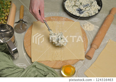 Woman's hand with fork putting cottage cheese with parsley and dill greens on dough. Rolling pin, cutter, egg, green towel and microgreens sprouts on kitchen table. Home cooking baking puffs 124316463