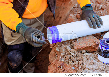 Construction worker prepares PVC piping by applying purple primer to ensure durable seal in trench setting. 124320753