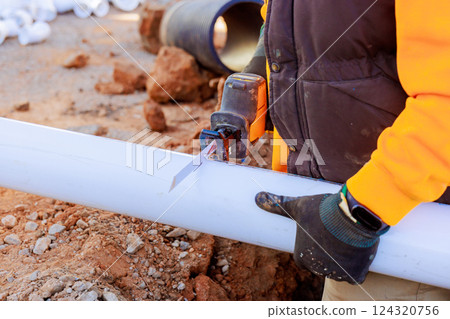 Plumber in yellow jacket uses tool to cut white pipe at construction site surrounded by dirt 124320756