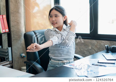 A young girl takes a break to stretch at her office desk, promoting a healthy lifestyle in a modern workspace. A young girl takes a break to stretch at her office desk, promoting a healthy lifestyle in a modern workspace. 124320764