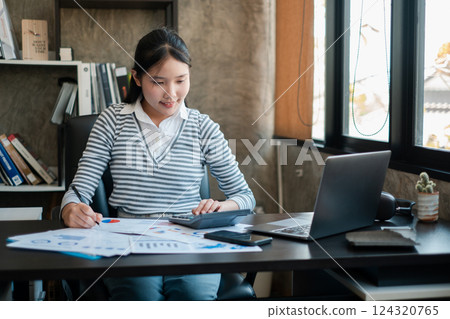 A young woman working at a desk, analyzing financial documents with a laptop in a modern office setting. 124320765