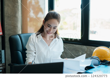 Smiling woman in a modern office setting, reviewing architectural plans with safety gear and headphones on the desk. 124320773