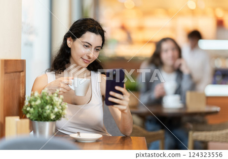 Woman holding mobile phone in her hands and drinking coffee while sitting at a table 124323556