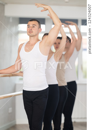 Experienced young male ballet dancer, dressed in white and black and group of girls and guys engaged in classical ballet performs exercise standing in 3rd position near ballet barre in dance hall 124324007