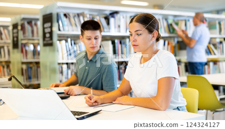 Positive students using laptop and writing in notepads in the book space library Positive students using laptop and writing in notepads in the book space library 124324127