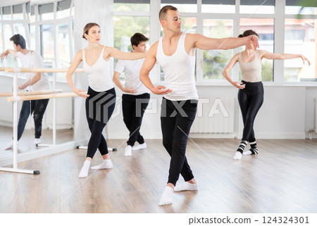 Experienced young male ballet dancer, dressed in white and black executing dance moves with focused precision among group of people rehearsing in ballet studio Experienced young male ballet dancer, dressed in white and black executing dance moves with focused precision among group of people rehearsing in ballet studio 124324301