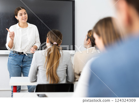Young female teacher near interactive board, gesturing with hand in front of people sitting at desks in auditory Young female teacher near interactive board, gesturing with hand in front of people sitting at desks in auditory 124324309