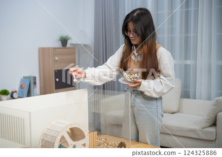 Woman Feeding Hamster in a Modern Living Room with a Transparent Cage and Cozy Interior Design Elements Emphasizing Pet Care and Comfort Woman Feeding Hamster in a Modern Living Room with a Transparent Cage and Cozy Interior Design Elements Emphasizing Pet Care and Comfort 124325886