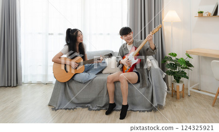 Two Asian Friends Playing Acoustic Guitar and Electric Guitar Together in a Bright Living Room Setting with Natural Light Streaming Through Large Windows Two Asian Friends Playing Acoustic Guitar and Electric Guitar Together in a Bright Living Room Setting with Natural Light Streaming Through Large Windows 124325921