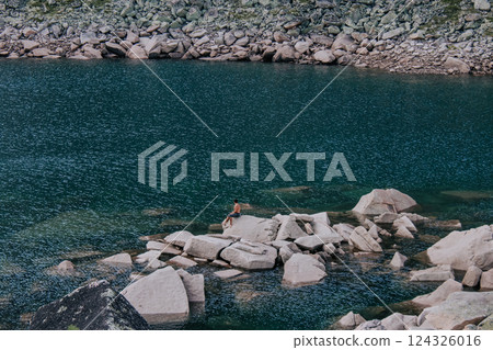 Man sits on large stone protruding from water near shore of emerald-colored mountain lake. Natural landscape, summer hiking day. Hike at kuturchinskoye belogorye Krasnoyarsk region. Tourism in Siberia Man sits on large stone protruding from water near shore of emerald-colored mountain lake. Natural landscape, summer hiking day. Hike at kuturchinskoye belogorye Krasnoyarsk region. Tourism in Siberia 124326016