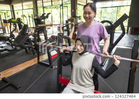 Arab middle eastern muslim woman on bench press training with chinese east asian personal trainer woman at the gym. Fitness exercise and healthy lifestyle concept 124327464